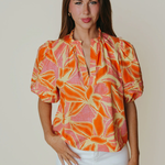 Woman wearing a colorful patterned blouse with white pants against a plain background