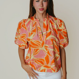 Woman wearing a colorful patterned blouse with white pants against a plain background