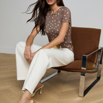 Woman sitting on a brown chair wearing a polka dot top and white pants in a minimalistic setting.