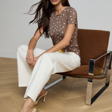 Woman sitting on a brown chair wearing a polka dot top and white pants in a minimalistic setting.