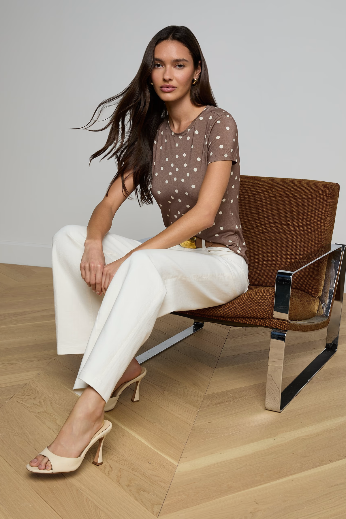 Woman sitting on a brown chair wearing a polka dot top and white pants in a minimalistic setting.