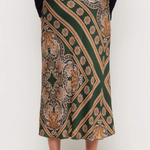 A model is shown from the waist down wearing a dark green midi skirt with a detailed gold, beige, and brown paisley pattern, paired with a black long-sleeve top and black high heels.