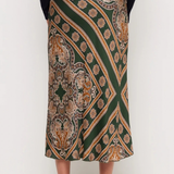A model is shown from the waist down wearing a dark green midi skirt with a detailed gold, beige, and brown paisley pattern, paired with a black long-sleeve top and black high heels.