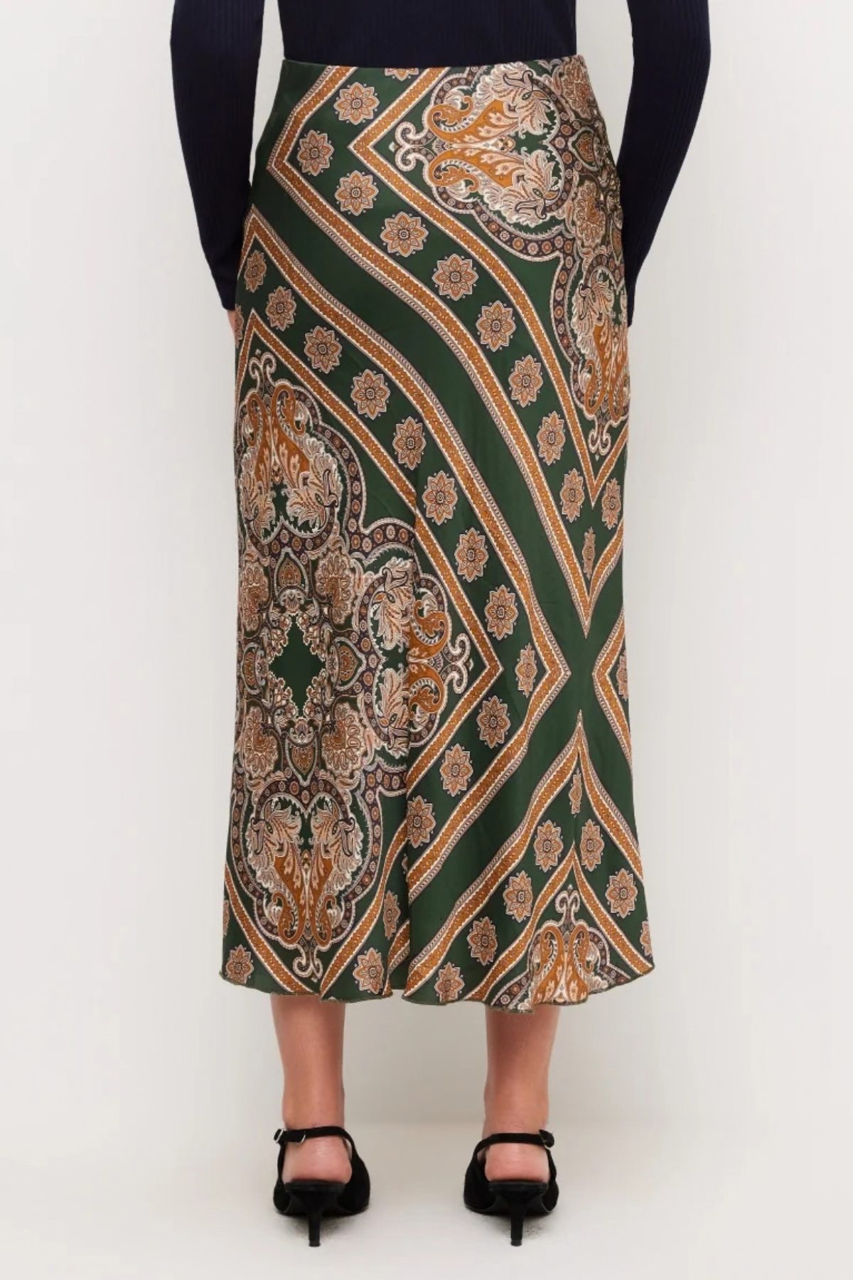 A model is shown from the waist down wearing a dark green midi skirt with a detailed gold, beige, and brown paisley pattern, paired with a black long-sleeve top and black high heels.