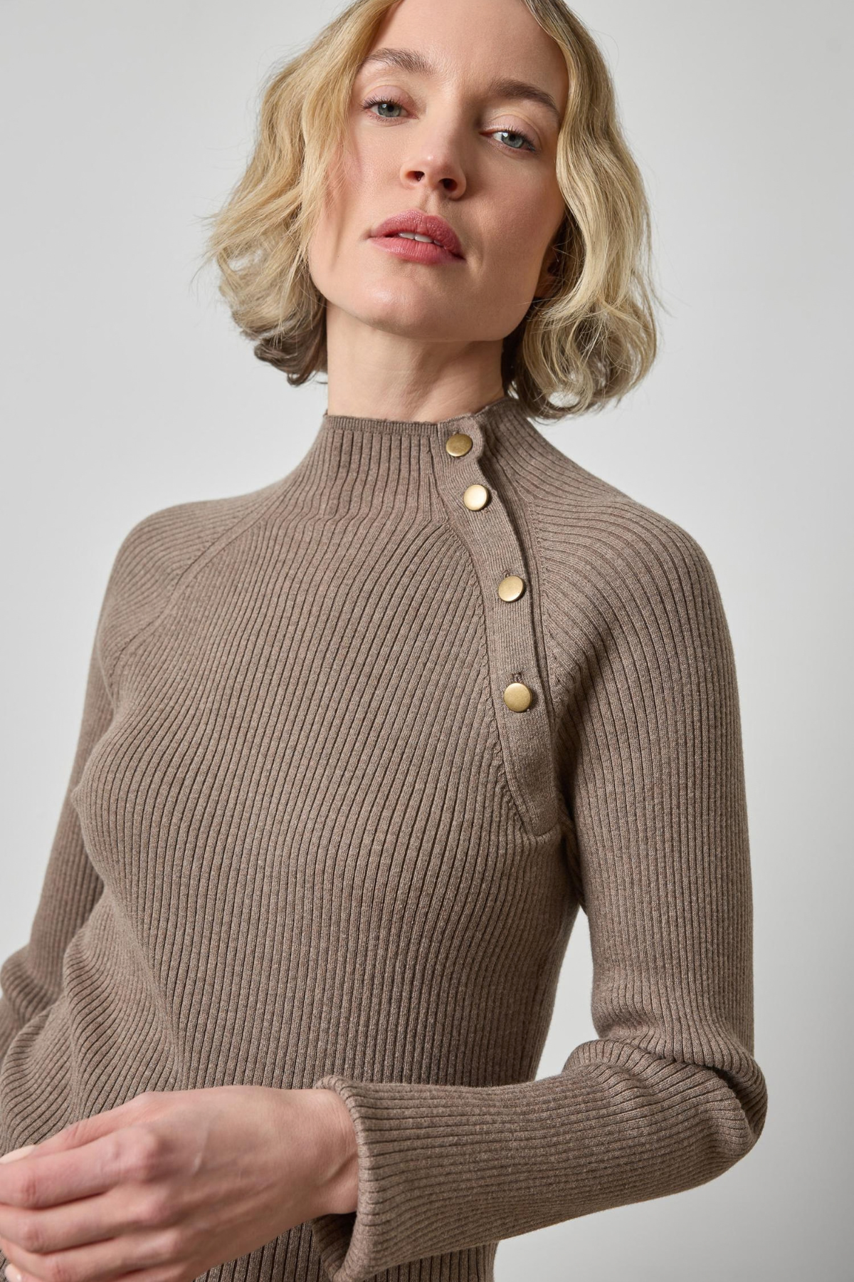 Woman wearing a brown ribbed sweater with gold buttons on a light gray background