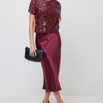 A woman models a two-piece outfit consisting of a short-sleeve burgundy sequined top and a matching satin midi skirt, holding a black clutch bag.