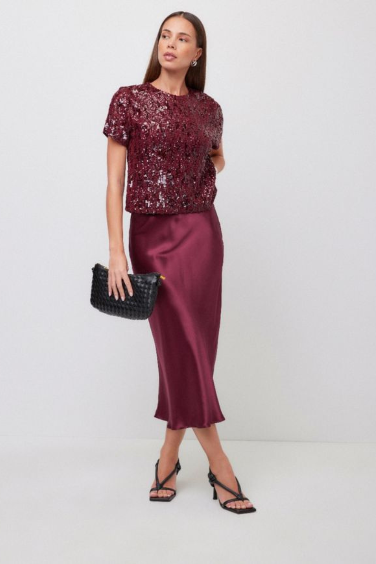 A woman models a two-piece outfit consisting of a short-sleeve burgundy sequined top and a matching satin midi skirt, holding a black clutch bag.