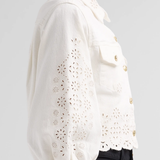 A side-profile view of a model wearing a white eyelet cropped jacket with gold buttons, paired with black jeans, against a light gray background.