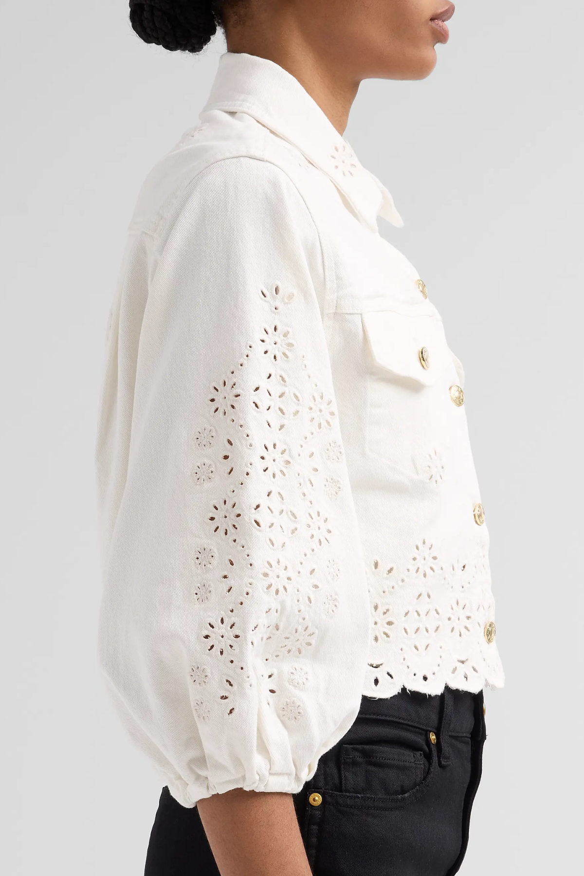 A side-profile view of a model wearing a white eyelet cropped jacket with gold buttons, paired with black jeans, against a light gray background.
