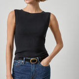 A woman wearing a sleeveless black ribbed knit top with a boat neckline, paired with blue jeans and a black belt with a gold buckle, against a light gray studio background.