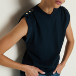 Woman wearing a dark blue sleeveless top with button details and blue jeans against a plain background