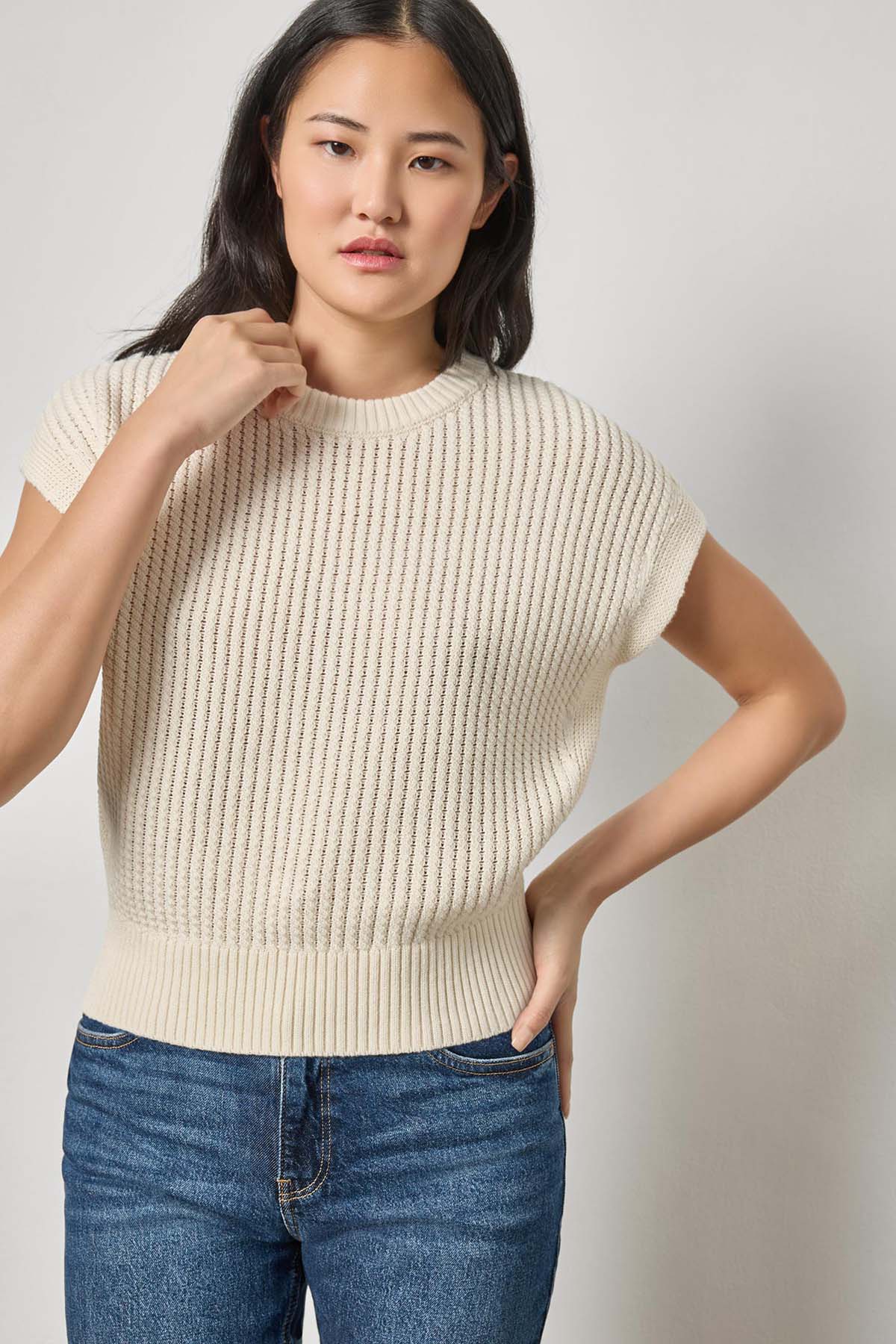 Woman wearing a cream ribbed sweater and blue jeans against a plain background