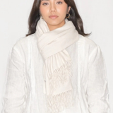 A woman wearing a light cream-colored scarf with fringe detailing and a matching white textured top, photographed against a plain white background.