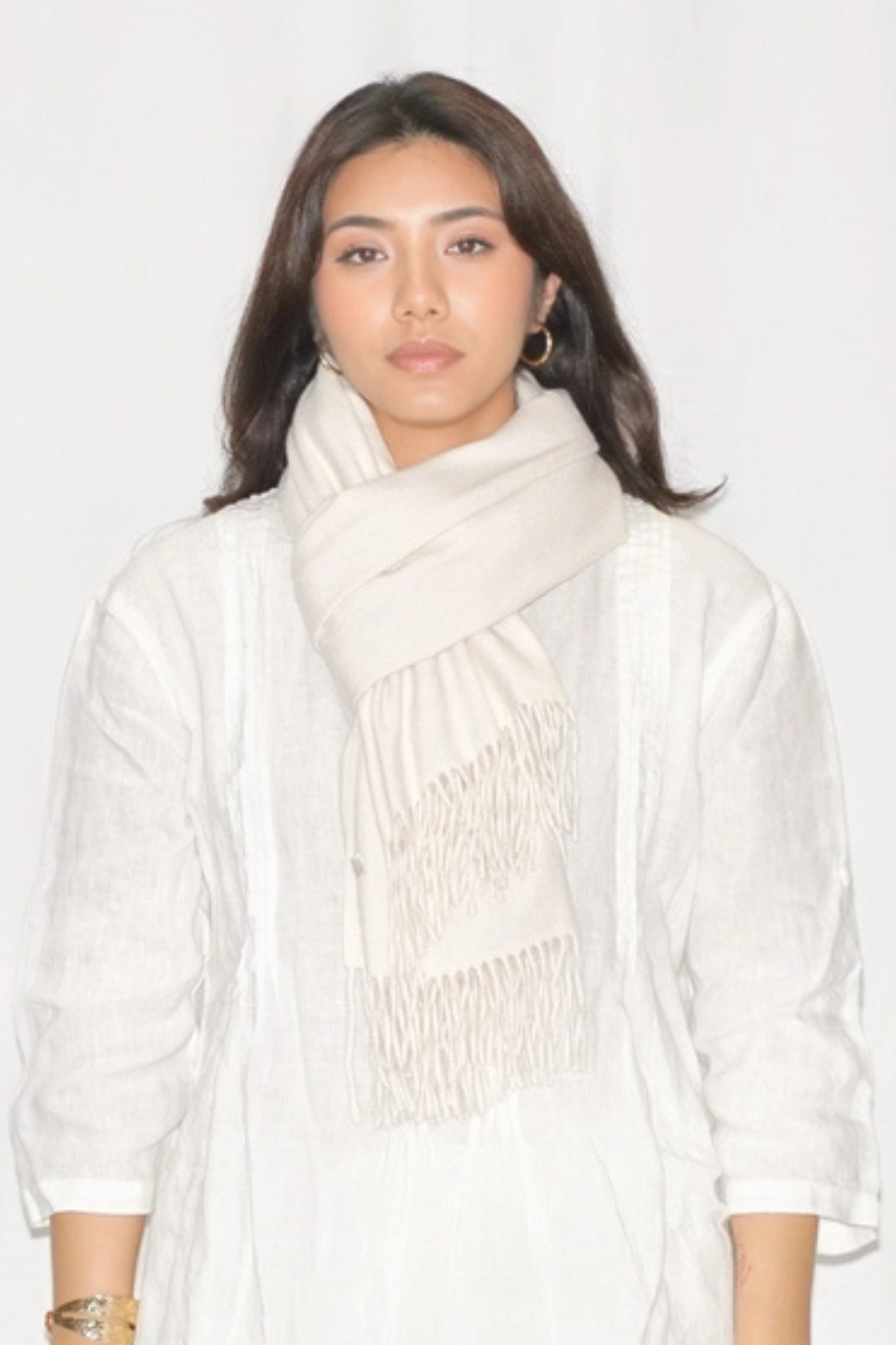 A woman wearing a light cream-colored scarf with fringe detailing and a matching white textured top, photographed against a plain white background.
