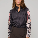 A woman modeling a dark charcoal gray satin button-up blouse with long sleeves featuring a pink and white floral print, paired with dark brown high-waisted trousers with button details on the front.