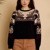 A woman modeling a black and beige knit sweater with intricate floral and geometric patterns on the yoke and sleeves, paired with brown pants, set against a warm beige background.