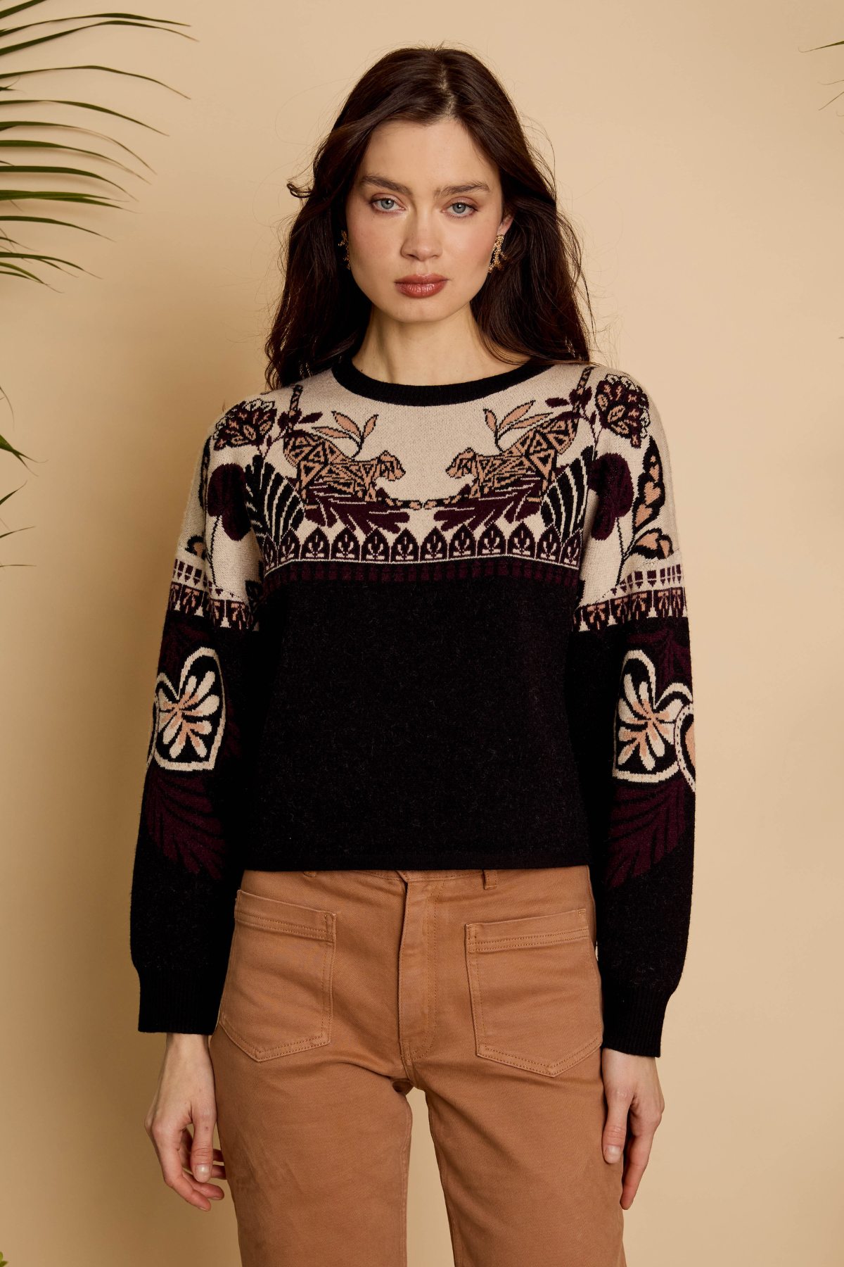 A woman modeling a black and beige knit sweater with intricate floral and geometric patterns on the yoke and sleeves, paired with brown pants, set against a warm beige background.