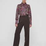 A model stands against a light gray studio background wearing a long-sleeve high-neck blouse with a purple, pink, and green paisley pattern, paired with dark brown wide-leg trousers and black pointed shoes.