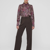 A model stands against a light gray studio background wearing a long-sleeve high-neck blouse with a purple, pink, and green paisley pattern, paired with dark brown wide-leg trousers and black pointed shoes.