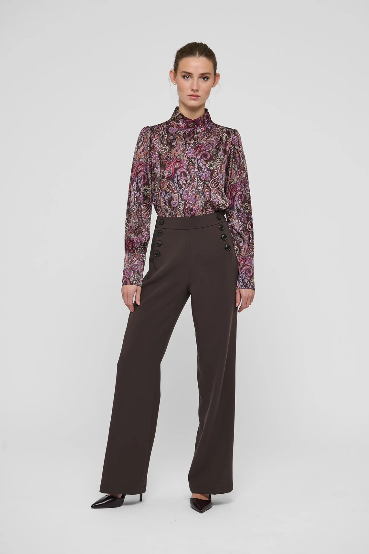 A model stands against a light gray studio background wearing a long-sleeve high-neck blouse with a purple, pink, and green paisley pattern, paired with dark brown wide-leg trousers and black pointed shoes.