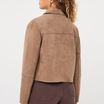 A woman shown from the back wearing a cropped taupe-brown suede jacket and dark brown pants, photographed against a clean light gray studio background.