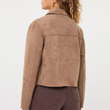 A woman shown from the back wearing a cropped taupe-brown suede jacket and dark brown pants, photographed against a clean light gray studio background.
