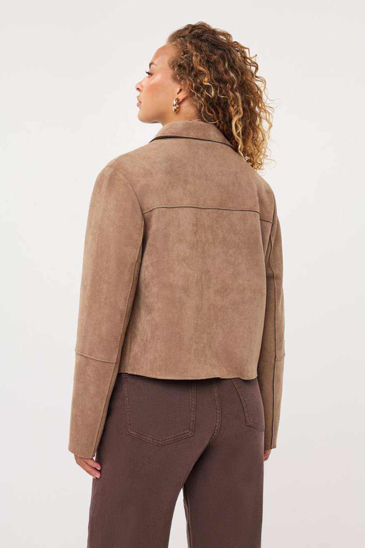 A woman shown from the back wearing a cropped taupe-brown suede jacket and dark brown pants, photographed against a clean light gray studio background.