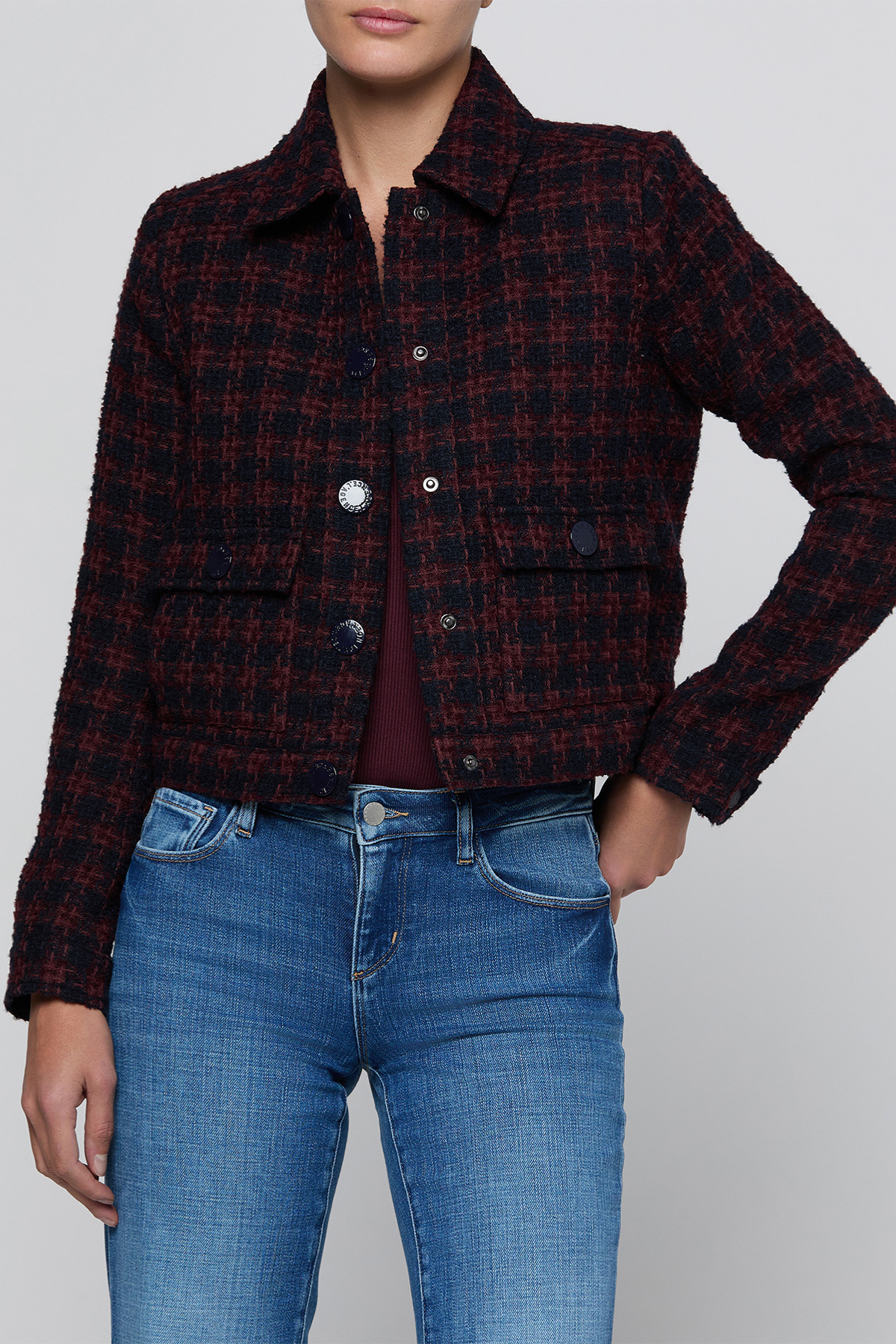A cropped, long-sleeve plaid jacket in dark red and black tweed, worn by a model with blue jeans against a light gray studio background.