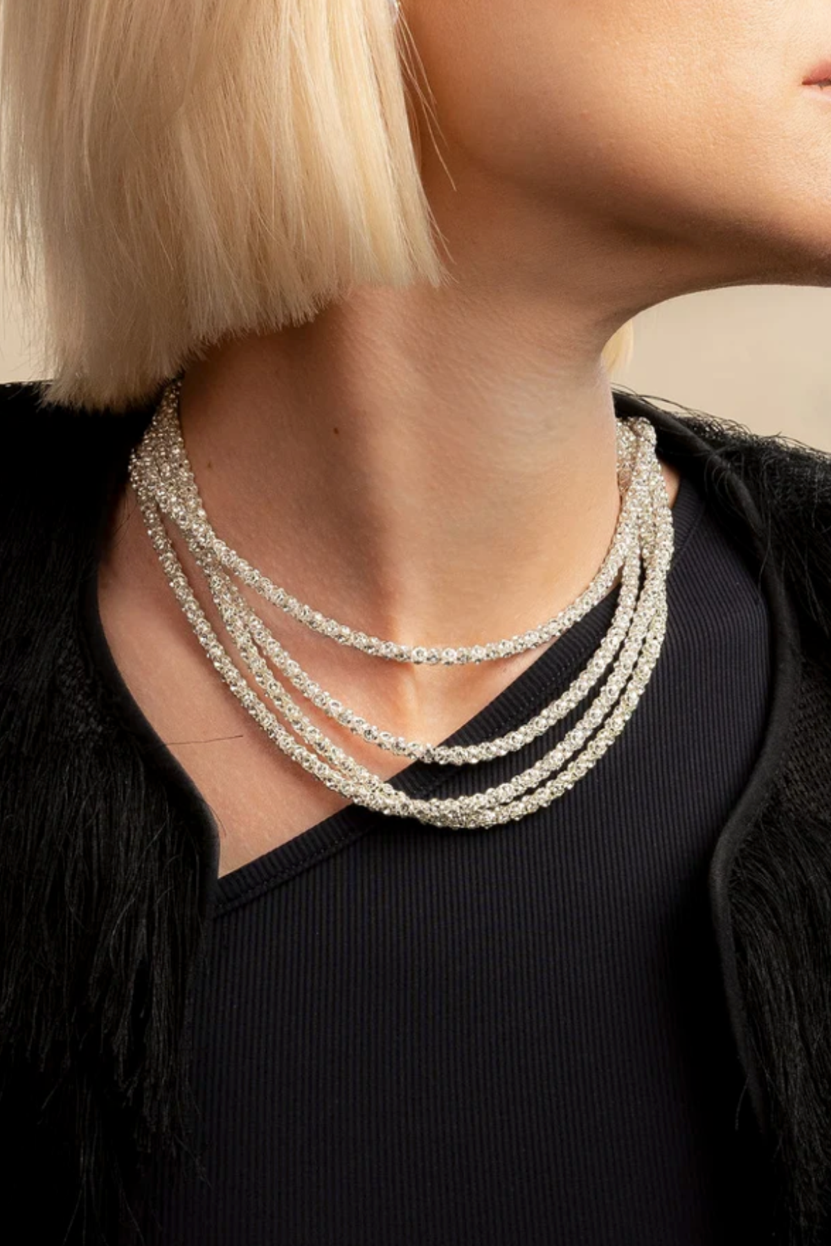 A close-up of a model wearing a multi-strand pearl necklace with a textured floral pattern, styled with a black top and black fringed jacket.