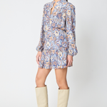 A woman modeling a long-sleeve mini dress with a blue, orange, and white paisley pattern, paired with beige knee-high boots, against a light gray studio background.