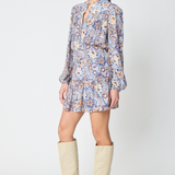A woman modeling a long-sleeve mini dress with a blue, orange, and white paisley pattern, paired with beige knee-high boots, against a light gray studio background.