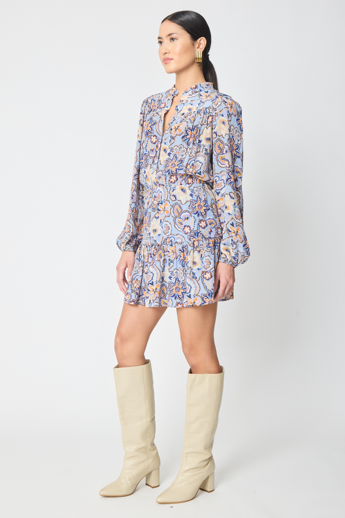 A woman modeling a long-sleeve mini dress with a blue, orange, and white paisley pattern, paired with beige knee-high boots, against a light gray studio background.