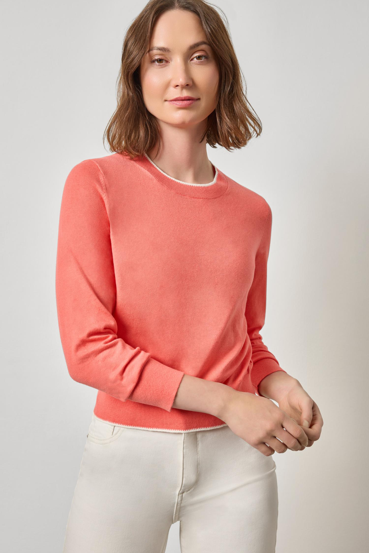 Woman wearing a coral sweater and white pants against a light gray background
