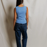 Person wearing a blue sleeveless top and dark jeans standing against a plain white background