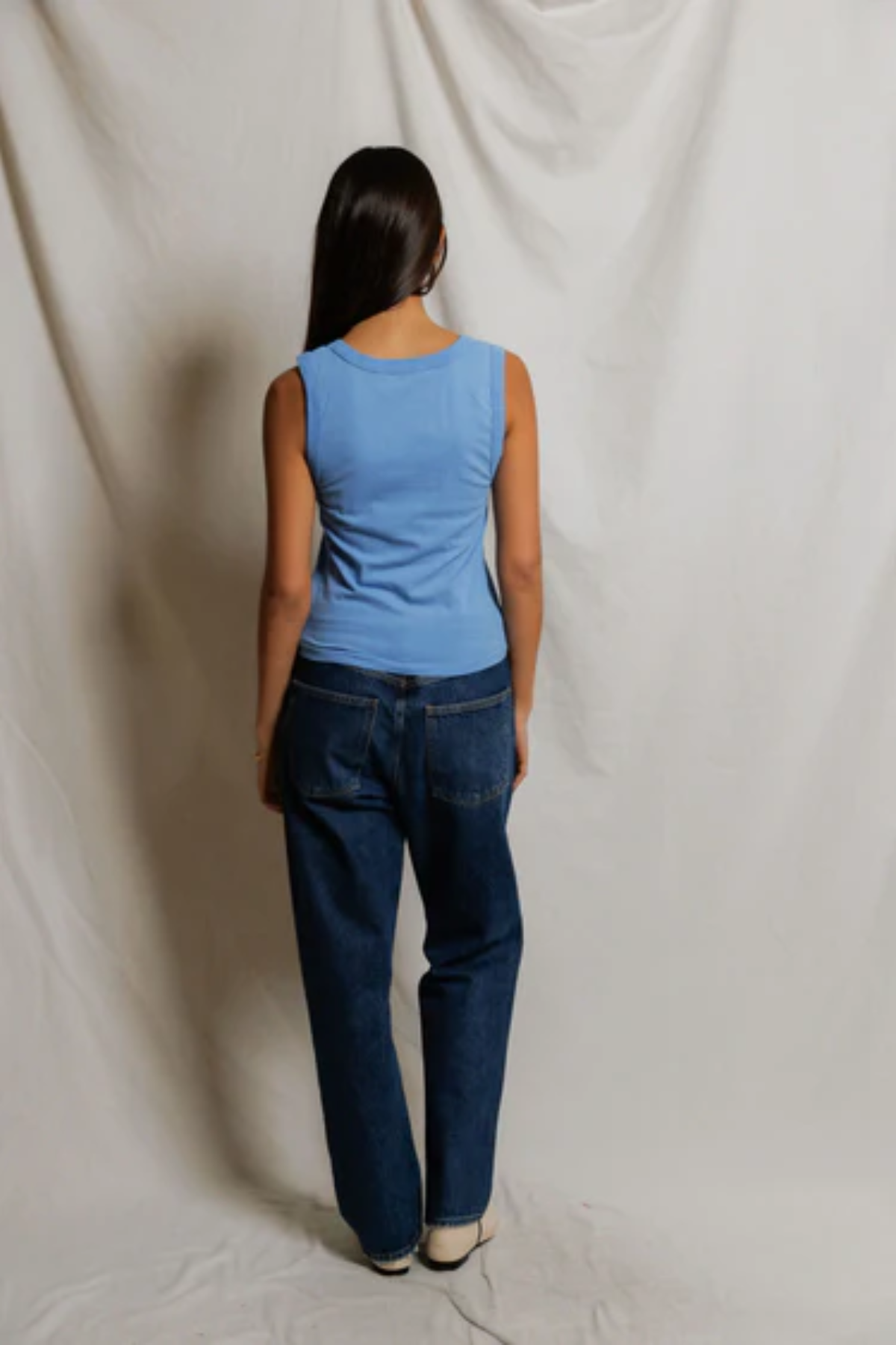 Person wearing a blue sleeveless top and dark jeans standing against a plain white background