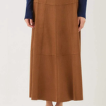 A model is wearing a brown, high-waisted, midi-length pleated skirt with a structured, paneled design, styled with a navy blue long-sleeve top and black pointed-toe flats against a plain white studio background.