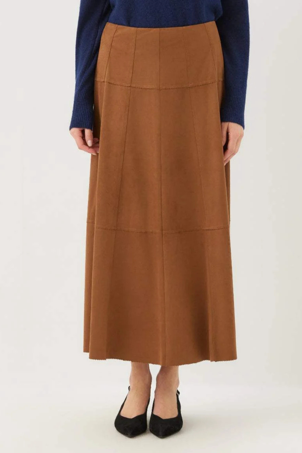 A model is wearing a brown, high-waisted, midi-length pleated skirt with a structured, paneled design, styled with a navy blue long-sleeve top and black pointed-toe flats against a plain white studio background.