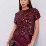 A woman modeling a short-sleeve burgundy sequined top paired with a matching satin skirt, holding a small black clutch against a light gray studio background.