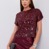 A woman modeling a short-sleeve burgundy sequined top paired with a matching satin skirt, holding a small black clutch against a light gray studio background.