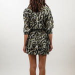 A woman viewed from behind wearing a short-sleeve mini dress with a black, olive green, and beige abstract pattern, standing against a plain white studio background.
