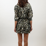 A woman viewed from behind wearing a short-sleeve mini dress with a black, olive green, and beige abstract pattern, standing against a plain white studio background.