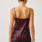 A woman shown from the back wearing a burgundy sequined camisole top with thin straps and black pants, against a plain light gray studio background.