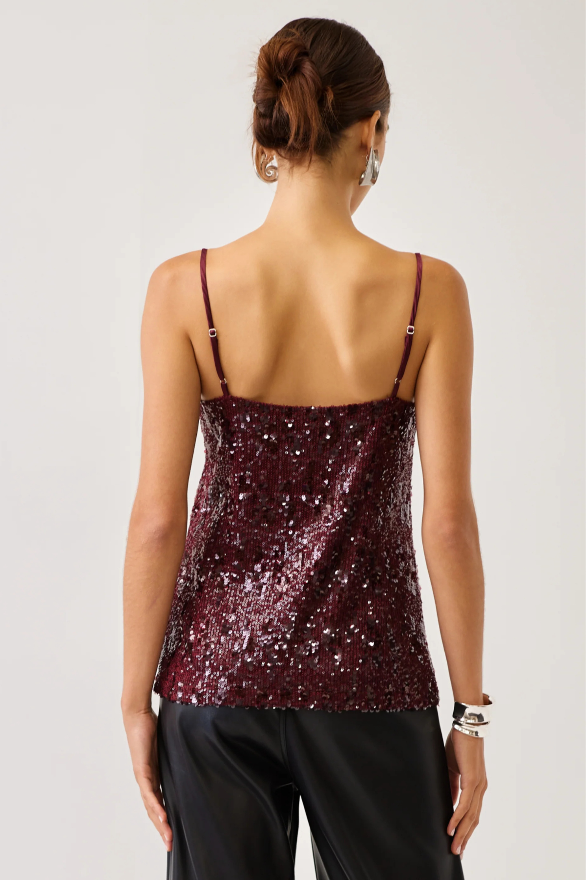 A woman shown from the back wearing a burgundy sequined camisole top with thin straps and black pants, against a plain light gray studio background.