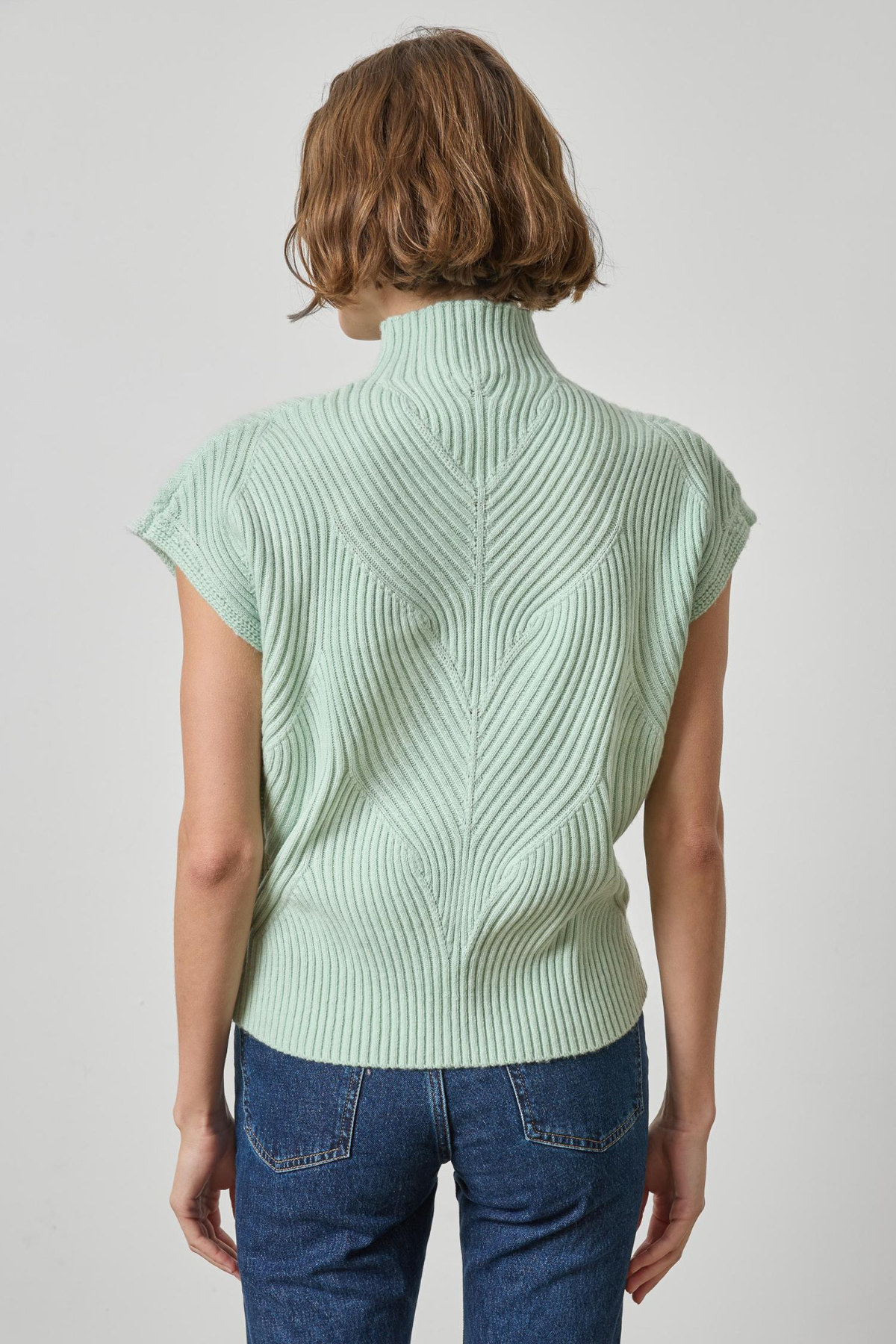 A woman viewed from the back wearing a pale mint green ribbed knit mock-neck top with short sleeves and blue jeans, against a light gray studio background.