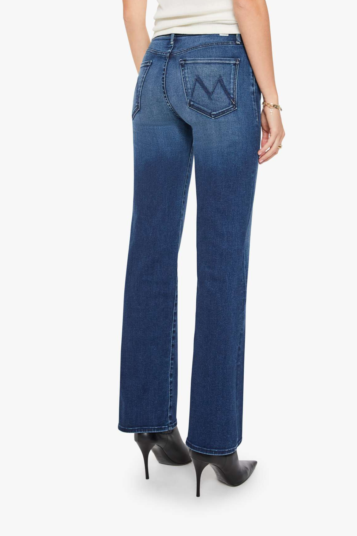 Rear view of a model wearing blue denim jeans with a distinctive 'M' logo on the back pocket, paired with black high-heeled boots against a light gray background.