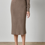 A model wearing a taupe brown ribbed knit midi skirt and matching knit top, paired with beige strappy heels, against a neutral studio backdrop.