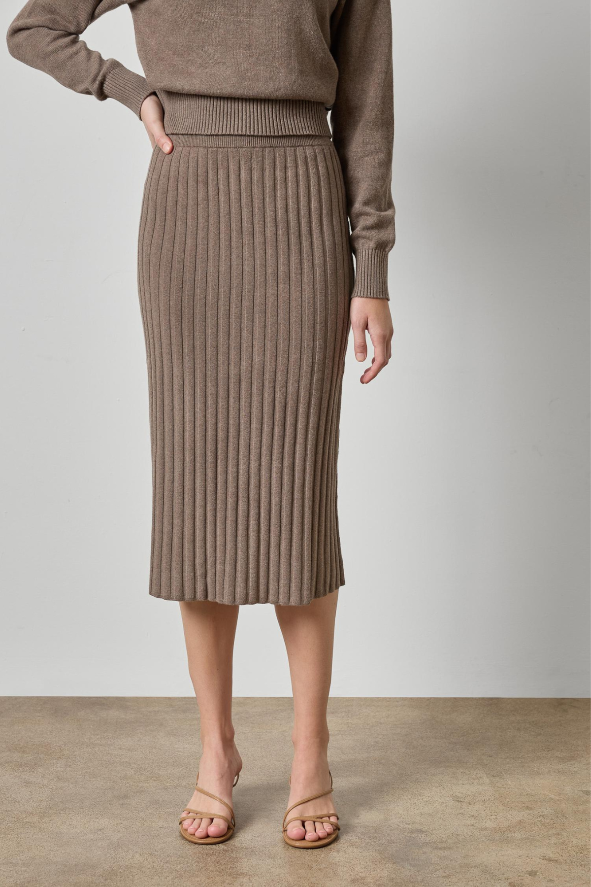A model wearing a taupe brown ribbed knit midi skirt and matching knit top, paired with beige strappy heels, against a neutral studio backdrop.