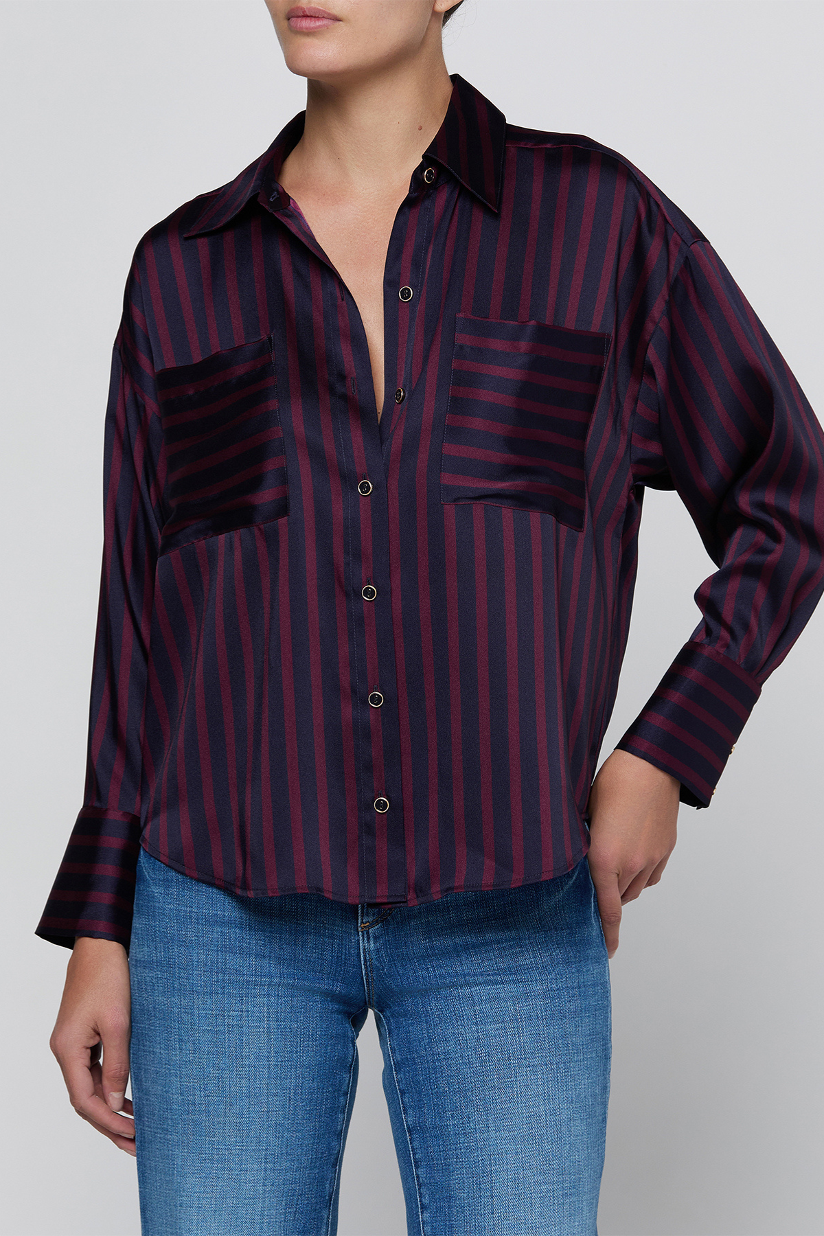 A woman wearing a dark striped button-up shirt with dark pockets and blue jeans, photographed against a light gray studio background.
