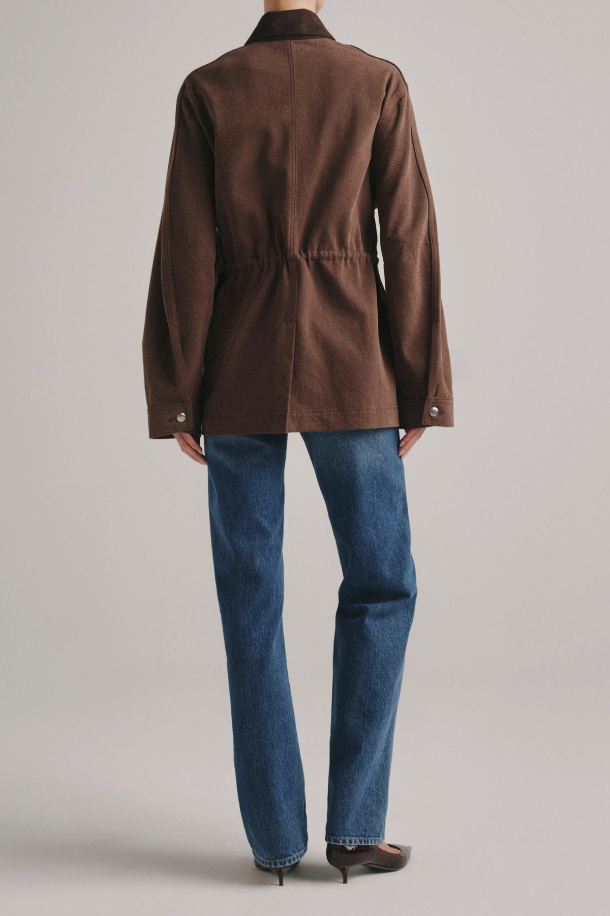 A model shown from the back wearing a brown corduroy jacket with a drawstring waist and blue jeans, against a light gray studio background.