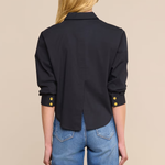 A woman shown from the back wearing a black long-sleeve button-up shirt with gold button accents at the cuffs, paired with blue jeans, against a solid beige background.